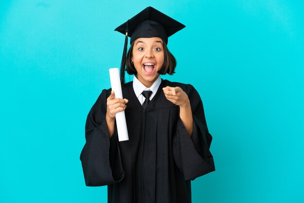 Jeune fille diplômée de l'université sur fond bleu isolé surpris et pointant vers l'avant