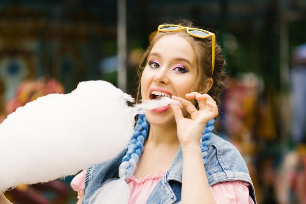 Une jeune fille drôle mange du coton sucré par une journée ensoleillée dans un parc d'attractions.