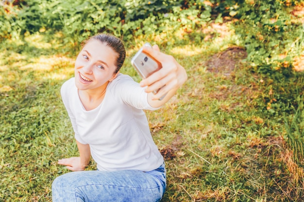 Jeune fille drôle prendre selfie des mains avec téléphone assis sur le parc d'herbe verte ou le jardin. Portrait de jeune femme séduisante faisant selfie photo sur smartphone en journée d'été.