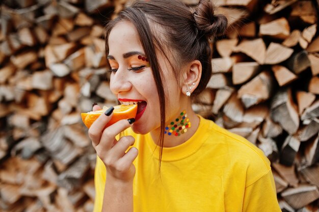 Jeune fille drôle avec des vêtements de maquillage lumineux sur une chemise jaune mange des morceaux d'orange sur fond de bois