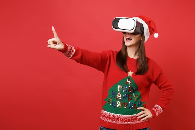 Jeune fille du Père Noël regardant dans le casque, touche quelque chose comme un clic sur le bouton, pointant sur un écran virtuel flottant isolé sur fond rouge. Bonne année 2019 concept de fête de vacances célébration.