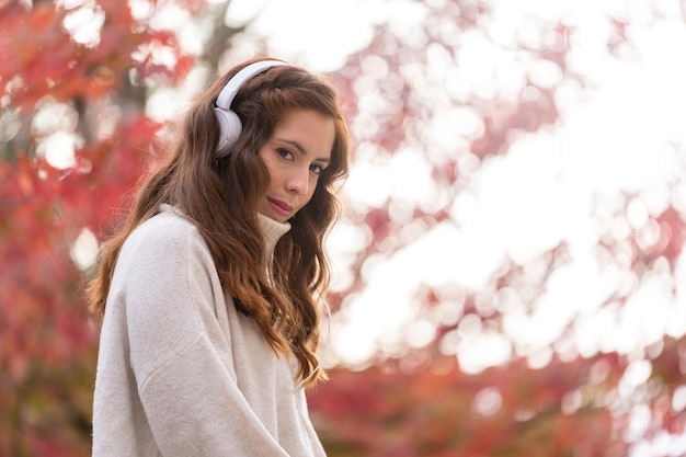 Jeune fille écoutant de la musique à l'extérieur avec un casque blanc avec des arbres feuillus rouges en arrière-plan