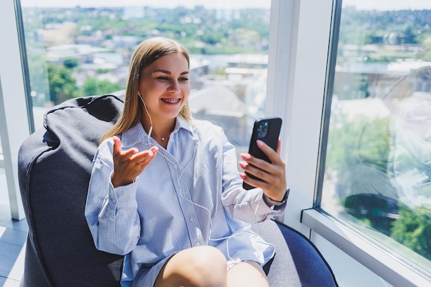 Une jeune fille écoute de la musique sur des écouteurs et regarde quelque chose sur un téléphone mobile Concept de style de vie de femme moderne Jeune femme assise dans un espace de coworking intérieur de bureau