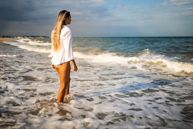 Une jeune fille élancée dans un maillot de bain bleu doux et une chemise blanche, se promène le long d'une large plage de sable près de la mer bleue avec des vagues blanches