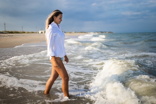 Une jeune fille élancée dans un maillot de bain bleu doux et une chemise blanche, se promène le long d'une large plage de sable près de la mer bleue avec des vagues blanches