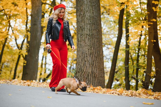 Une jeune fille élégante aux longs cheveux clairs dans des lunettes ensoleillées se promène avec un petit chien moyen un carlin par le bouledogue français dans un parc au printemps en automne