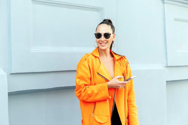 Jeune fille élégante dans une rue de la ville avec un téléphone Belle brune dans une veste orange vif et des lunettes de soleil La vie moderne de la jeunesse