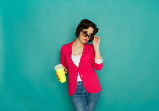 Jeune fille élégante dans des vêtements décontractés lumineux et des lunettes de soleil avec boisson à emporter. Jeune femme observatrice posant sur fond de studio azur, espace pour copie