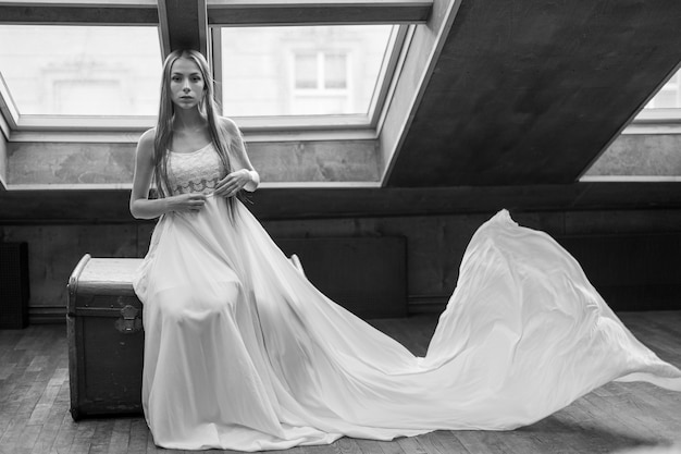 Photo jeune fille élégante romantique en longue robe flottante blanche assise sur la poitrine de la dot dans le loft