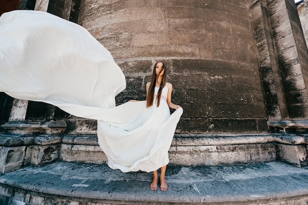 Jeune fille élégante romantique en longue robe fluide blanche posant sur mur ancien en pierre