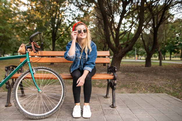 Jeune fille élégante souriante assise sur un banc dans un parc avec un vélo et au repos.