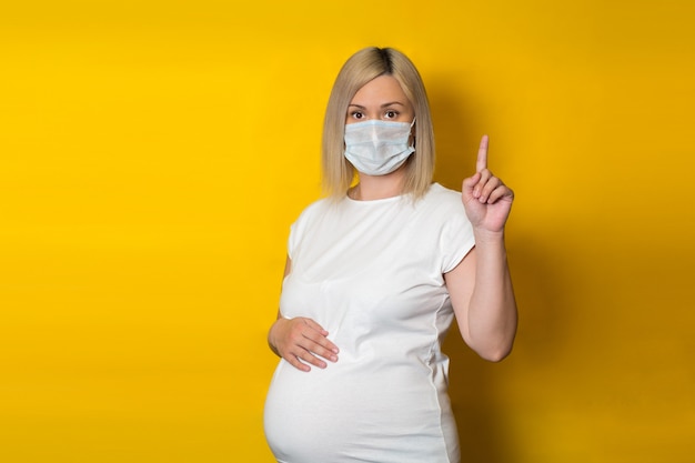 Photo une jeune fille enceinte dans un masque de protection tenant son doigt vers le haut et avec sa main sur son ventre. concept de soins de santé