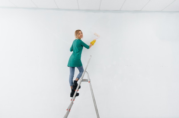 Une jeune fille sur un escabeau peint un mur blanc avec un rouleau. Réparation de l'intérieur.