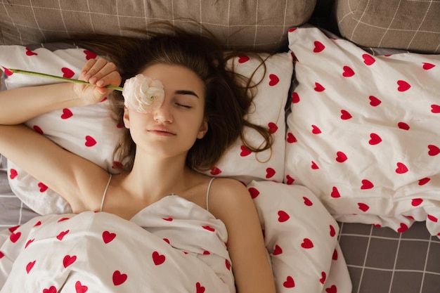 La jeune fille est allongée dans son lit avec des coeurs rouges et tient une fleur dans sa main.