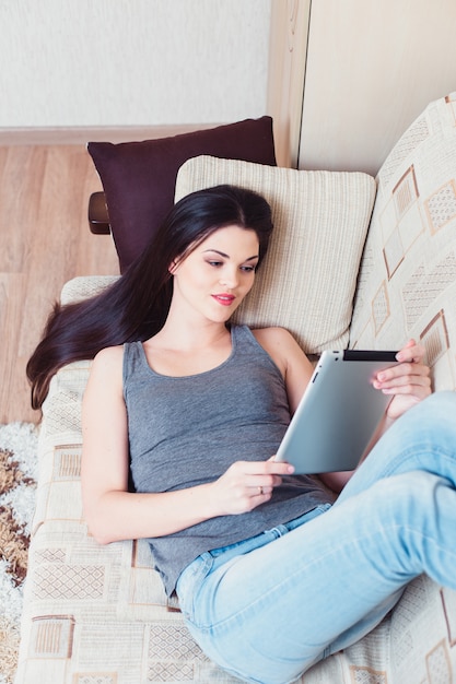 La jeune fille est allongée avec une tablette sur un canapé beige