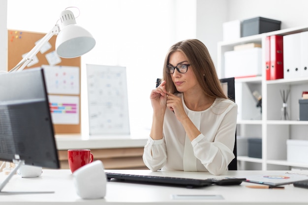 Une jeune fille est assise au bureau du bureau, tenant un crayon à la main et regardant l’écran.