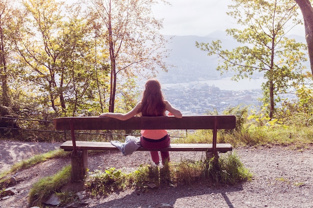 La jeune fille est assise sur un banc dans la forêt, dos à la caméra et devant elle s'ouvre un panorama de la ville