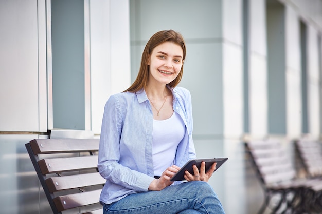 Une jeune fille est assise sur un banc avec une tablette dans les mains et sourit.