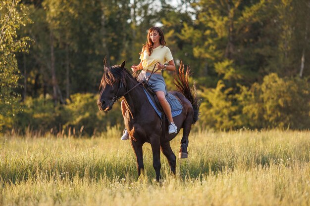 Une jeune fille est assise sur un cheval sur fond de forêt au coucher du soleil.