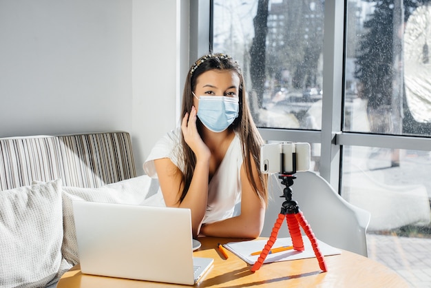 Une jeune fille est assise dans un café dans un masque et dirige un blog vidéo. Communication avec la caméra.