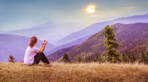 Une jeune fille est assise sur l'herbe avec des montagnes et regarde au coucher du soleil