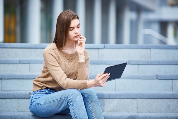 Une jeune fille est assise sur les marches et regarde la tablette.