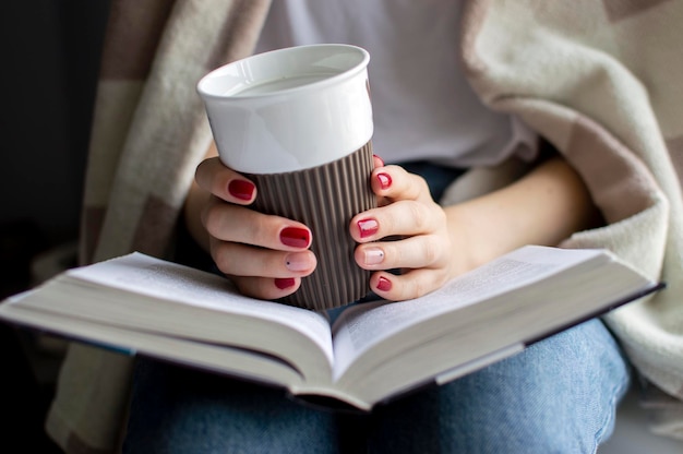 Jeune fille est assise près de la fenêtre recouverte d'une couverture, elle lit un livre et boit