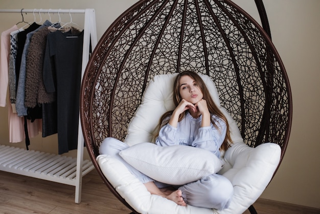 La jeune fille est assise en pyjama sur une chaise cocon. Intérieur élégant de la pièce. La fille flirte.