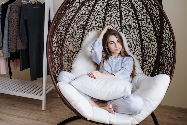 La jeune fille est assise en pyjama sur une chaise cocon. Intérieur élégant de la pièce.