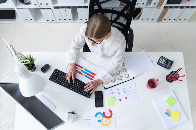 Une jeune fille est assise à une table dans le bureau, tenant un crayon à la main et tapant sur le clavier.