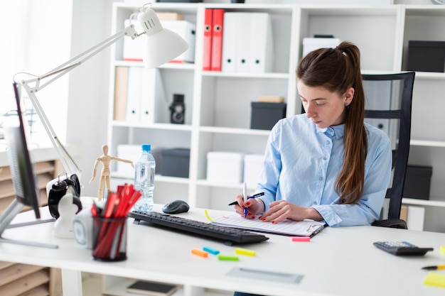 Une jeune fille est assise à une table dans le bureau, tenant un stylo à la main et travaillant avec la documentation.