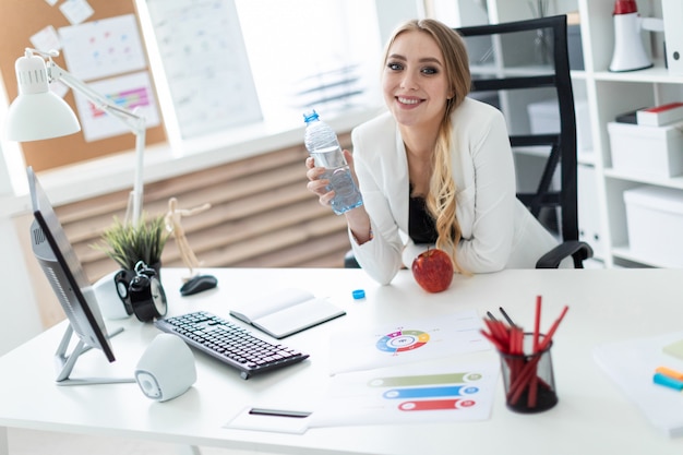 Une Jeune Fille Est Assise à Une Table Dans Le Bureau Et Tient Une Bouteille D’eau à La Main. Avant La Fille Sur La Table Est Une Pomme.