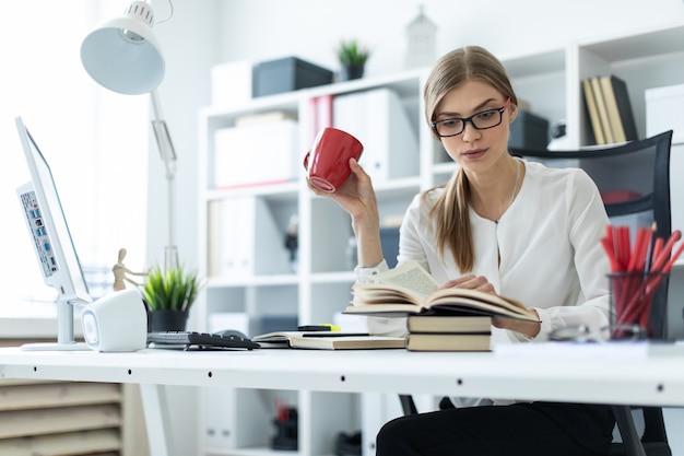 Une jeune fille est assise à une table dans le bureau et tient une tasse rouge à la main et lit un livre.