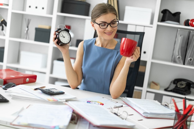 Une jeune fille est assise à une table dans son bureau, tenant un réveil et une tasse rouge.