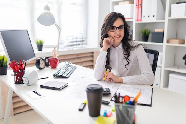 Une jeune fille est assise à la table du bureau et tient un stylo dans la main gauche.