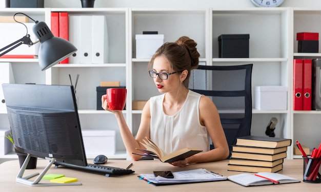 Une Jeune Fille Est Assise à Une Table D'ordinateur Et Tient Un Livre Ouvert Et Une Tasse Rouge Dans Ses Mains.