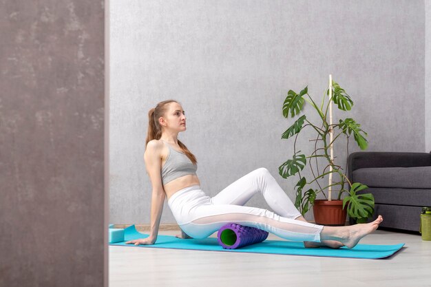 Une jeune fille est assise sur un tapis de yoga à la maison dans le salon et fait de l'automassage avec un rouleau myofascial
