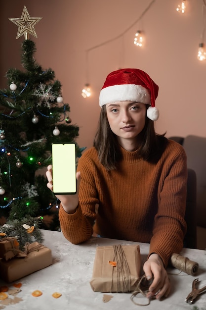 une jeune fille est vêtue d'un pull marron sur la tête un bonnet de noel dans les mains tient un téléphone portable