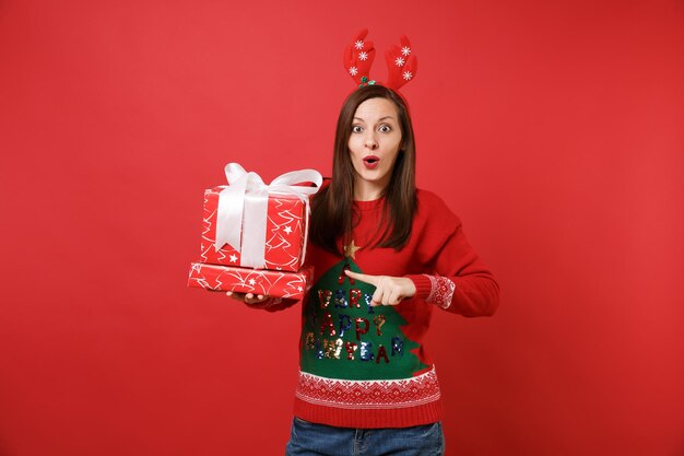 Jeune fille étonnée de Santa dans des cornes de cerf décoratives amusantes sur la tête, pointant l'index sur des boîtes rouges avec des cadeaux cadeaux isolés sur fond rouge. Bonne année 2019 concept de fête de vacances célébration.