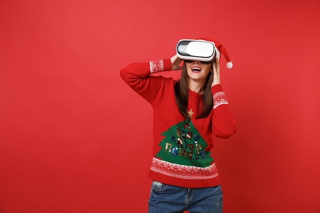 Jeune fille étonnée de Santa en pull tricoté chapeau de Noël regardant dans le casque mettant les mains sur la tête isolée sur fond rouge. Bonne année 2019 concept de fête de vacances célébration. Maquette de l'espace de copie.