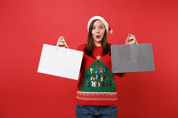 Jeune fille étonnée de Santa à la surprise, tenant des sacs de paquets avec des achats après le shopping isolé sur fond rouge. Bonne année 2019 concept de fête de vacances célébration. Maquette de l'espace de copie.