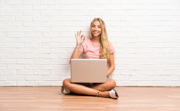 Jeune fille étudiante blonde avec un ordinateur portable sur le sol, montrant un signe ok avec les doigts