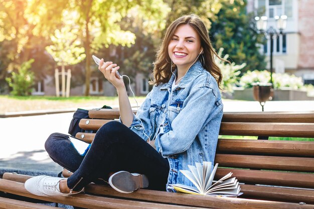 Jeune fille étudiante est assis sur un banc dans le parc et détient un téléphone portable. Fille écoute un livre audio dans le parc.