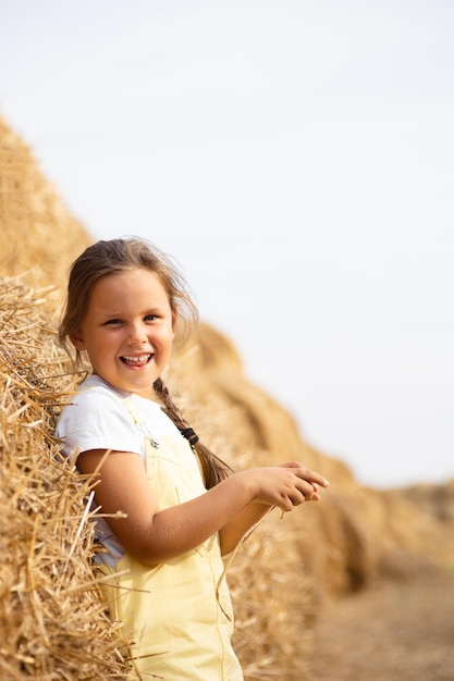 Jeune fille excitée avec une tresse montrant la langue souriante et regardant la caméra appuyée sur une botte de foin dans les mains du champ tenant du foin avec les yeux plissés Temps loin de la ville dans le champ de campagne