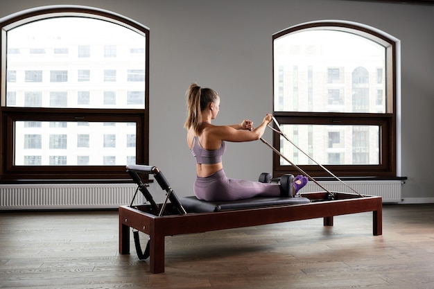 Jeune fille faisant des exercices de Pilates avec un lit réformateur. Bel entraîneur de fitness mince sur fond gris réformateur, discret, lumière d'art, bannière publicitaire de l'espace de copie