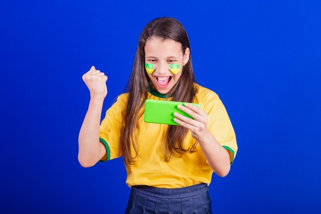 Jeune fille fan de football du Brésil tenant un téléphone portable regardant le jeu acclamant les applications Smartphone