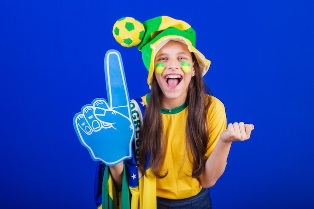 Jeune fille fan de football du Brésil vêtue d'un chapeau et d'un drapeau à l'aide d'un doigt en mousse faisant la fête incroyable