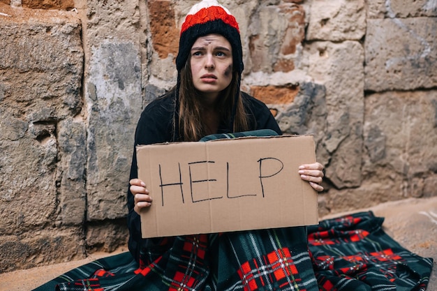 Une jeune fille ou une femme sans-abri déprimée assise seule sur une clôture sous un mur extérieur par temps froid, elle demande de l'aide
