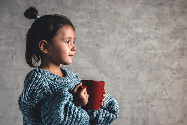 Jeune fille gaie portant des vêtements d'hiver et boire du chocolat chaud sur fond gris