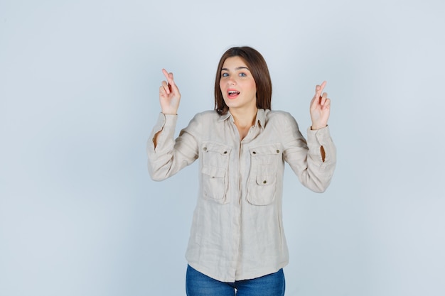 Jeune fille gardant les doigts croisés en chemise beige, jeans et l'air heureux. vue de face.